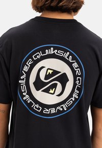 Camiseta de algodón negra con un diseño gráfico circular en la parte posterior, con el texto "Quiksilver" y un logotipo estilizado en acentos azules y amarillos.