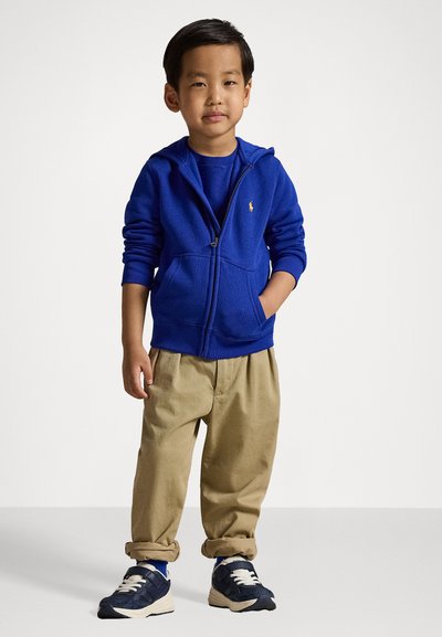 Μπλε φούτερ με φερμουάρ και τσέπες πάνω από μπλε πουκάμισο, συνδυασμένο με μπεζ παντελόνι διπλωμένα στους αστραγάλους και σκούρα αθλητικά παπούτσια.