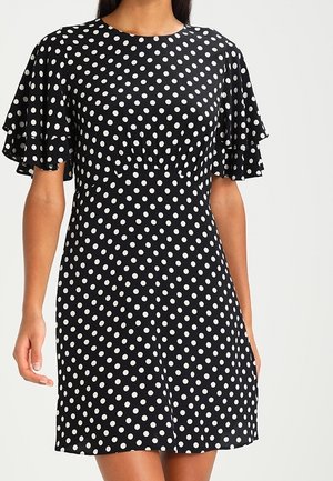 Schwarzes Kleid mit weißen Punkten, kurzen Flatterärmeln, tailliertem Schnitt und knielangem Saum an einer stehenden Person.