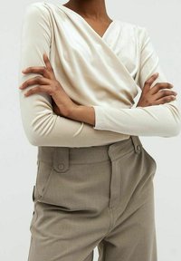 Světlé béžové zavinovací tričko s dlouhými rukávy, vyrobené z měkké, lesklé látky. Kombinované s vysokým pasem, světle šedé kalhoty s viditelnými knoflíky.