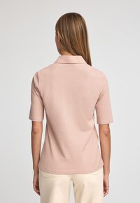 Jasnoróżowa koszulka polo z krótkim rękawem, z klasycznym kołnierzykiem i gładką, miękką teksturą. Widok z tyłu ukazuje dopasowaną sylwetkę.