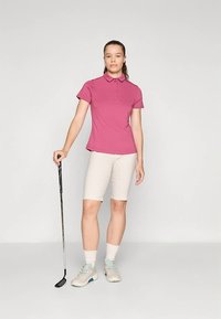 Rosa Poloshirt in figurbetontem Stil; kombiniert mit beigen Radshorts. Das Model hält einen Golfschläger, trägt hellblaue Sportschuhe und weiße Socken.