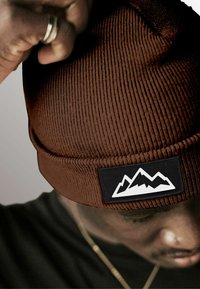 Brown ribbed beanie with a black patch featuring three white mountain silhouettes. Close-up shot focused on the hat's texture and details.