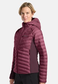 Donna che indossa una giacca a vento maroon trapuntata con cappuccio e pantaloni neri, in piedi contro uno sfondo chiaro e uniforme, rivolta in avanti con un leggero sorriso.