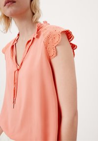 Blusa corallo a maniche corte con bordo in pizzo, collo con laccetto e tessuto morbido. Progettata con una vestibilità rilassata e senza motivi visibili.