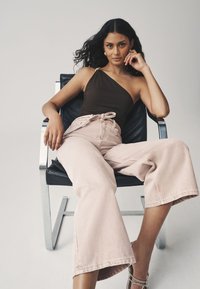 Barna egyrészes felső, világos rózsaszín, bő szárú nadrággal párosítva, megkötős derékkal és texturált anyagból, modern fekete széken ülve.