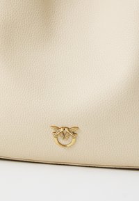 Béžová kožená kabelka s textúrovaným povrchom a zlatým logom s dvoma vtákmi. Dizajn je čistý a minimalistický.