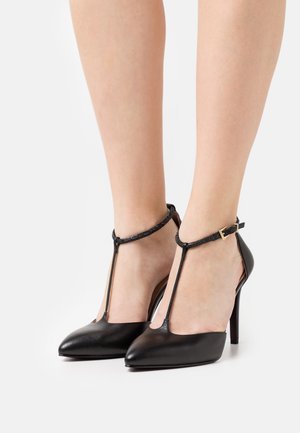 Chaussures à talons hauts en cuir noir avec un bout pointu, design T-strap et boucle de cheville. Présente une texture lisse et élégante.