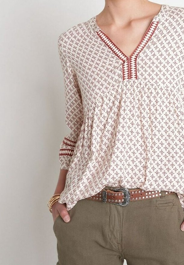 Blouse blanche avec des motifs géométriques bordeaux, col en V avec bordure brodée, manches longues, détail froncé à la taille, associée à un pantalon beige.