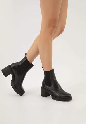 Bottes noires à la cheville en cuir avec un talon épais, des panneaux élastiques sur les côtés et une semelle texturée. Design minimaliste avec une languette pour un enfilage facile.