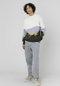 Pull avec motif de paysage montagneux en nuances de crème, vert et lavande ; poignets et ourlet côtelés. Associé à un jean gris clair et des baskets beige.