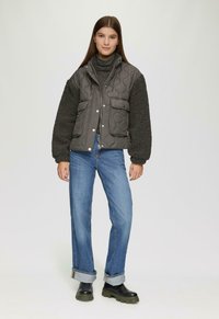 Jonge vrouw in een charcoal gewatteerde jas met fleece mouwen, grijze coltrui, opgerolde blauwe spijkerbroek en zwarte chunky laarzen, staand op een witte achtergrond.