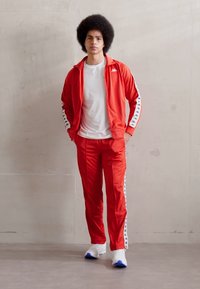 Survêtement rouge en matériau lisse, présentant des bandes latérales blanches avec un logo, associé à des chaussures blanches. Le modèle se tient contre un fond neutre.