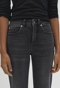 Person wearing black fitted jeans and a black long-sleeve shirt, hands relaxed by their sides against a plain background.