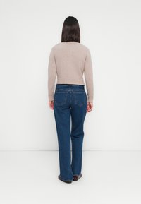 Vrouw met steil, donker haar, gekleed in een beige trui, blauwe rechte jeans en donkere schoenen, staand met haar gezicht naar een witte muur.