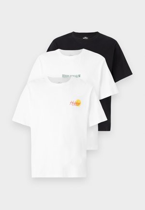 Tres camisetas de manga corta: una negra, una blanca con el texto "HIDDEN BUNGALOW", y una blanca con un gráfico de "Holidays" en rojo y amarillo. Tejido de algodón.