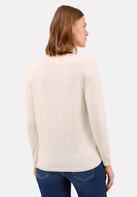 Donna con capelli castano chiaro fino alle spalle, vestita con un maglione lungo beige chiaro a maniche lunghe e jeans blu, voltata di spalle su uno sfondo neutro.