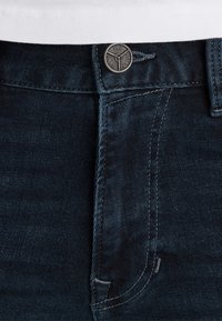 Tmavomodré džínsy s pásom a kovovou gombíkom s vygravírovaným nápisom "PME Legend Jeans" a časťou bielej košele nad nimi.