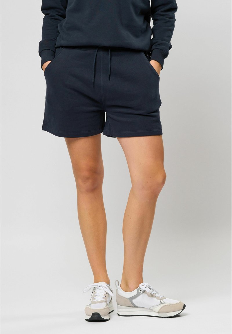Shorts en mélange de coton bleu marine avec une taille élastique et un cordon de serrage ; dotés de poches latérales et d'une coupe décontractée, associés à des baskets grises et blanches.