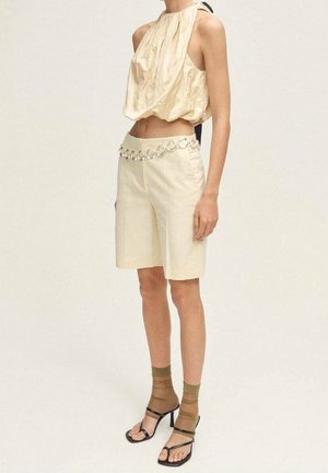 Femme portant un top sans manches crème à col halter, un short crème taille haute avec une ceinture chaîne argentée, des chaussettes marron transparentes et des sandales noires à talons.