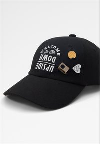 Melnā beisbola cepure ar baltu izšuvumu, kas saka "LAIPNI LŪDZAM UPSIDE DOWN" un trīs mazi dekoratīvi piespraudes sānos.