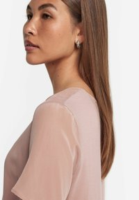 Ljusrosa blus med korta genomskinliga ärmar och rund halsringning. Slät tyg med subtil lyster, som visar på axelsömsdetaljer.