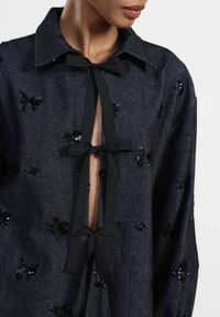 Top in denim di colore blu scuro con decorazioni in paillettes floreali nere. Presenta un colletto e un'apertura frontale fissata con lacci neri.
