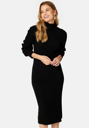 Vestito a coste nero con colletto alto, maniche lunghe e lunghezza fino al polpaccio. Presenta una fessura laterale per facilitare i movimenti.