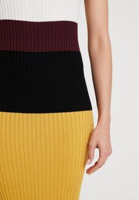 Knitklänning med ribbad struktur, med färgblock av vitt, vinrött, svart och gult. Närbild av midjan och handdetalj.