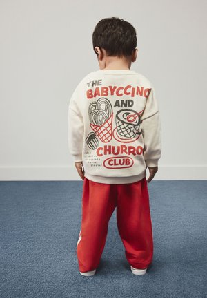 Kind mit dem Rücken zur Kamera trägt ein cremefarbenes Sweatshirt mit Lebensmittelmotiven und rote Hose, steht auf blauem Teppich vor weißer Wand.