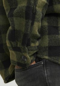 Groene en zwarte geruite flanellen shirt met een zachte textuur, met een zwarte knop op de manchetten en zwarte spijkerbroeken die gedeeltelijk zichtbaar zijn.