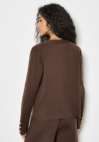 Maglione a maglia marrone con una vestibilità rilassata, caratterizzato da un collo rotondo e dettagli con bottoni sulle maniche. Tessuto testurizzato e finitura morbida.