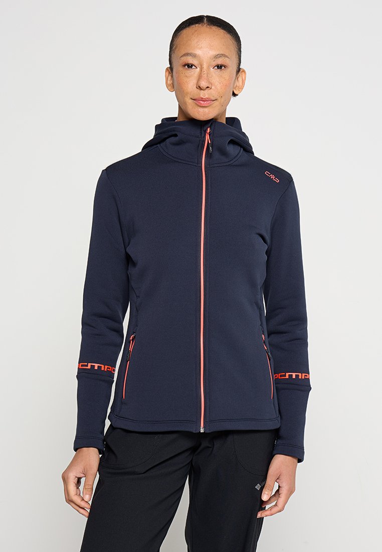 Femme portant un sweat à capuche bleu marine avec une fermeture éclair orange et des détails de logo, se tenant contre un fond clair uni.