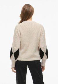 Světle růžový pletený svetr s texturovaným tělem, černé akcenty v diamantovém vzoru na objemových rukávech a žebrovaným lemem. Zadní pohled.