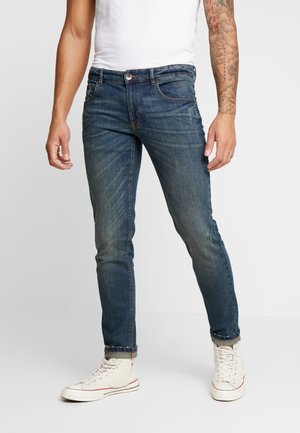 Man wearing fitted blue jeans, white sneakers, and a white sleeveless shirt, with a tattooed right arm, standing against a plain background.