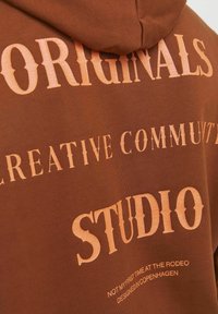 Bruine hoodie met grote, gestructureerde oranje tekst die "ORIGINALS", "CREATIVE COMMUNITY" en "STUDIO" zegt. Bevat kleinere tekst eronder.