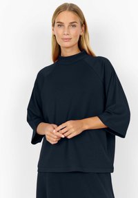 Tamno plavi oversized dukserica s visokim vratom i raglan rukavima, izrađena od mekanog materijala, s opuštenim krojem i minimalnim šavovima.