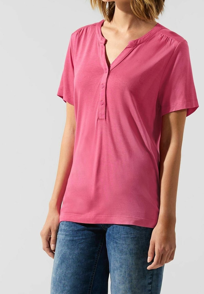 Maglietta henley rosa a maniche corte con chiusura a bottoni e delicati gather sulle spalle, realizzata in un tessuto leggero e liscio.
