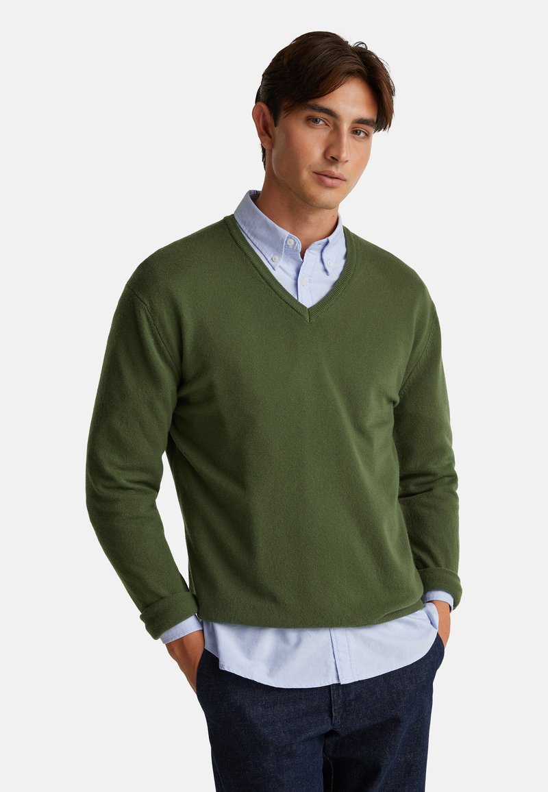 Zielony sweter w kształcie litery V z miękkiego materiału, z długimi rękawami, noszony na jasnoniebieskiej koszuli z kołnierzykiem, o luźnym kroju i minimalnych detalach.