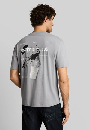 Mann mit lockigem Haar, der ein graues T-Shirt mit einer Kaffeetassen-Grafik und dem Text "Coffee Run Club" auf dem Rücken trägt.