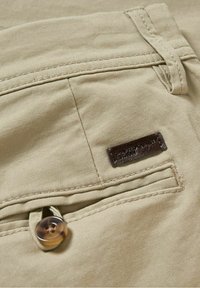Pantalon en tissu beige avec une poche arrière boutonnée et une petite étiquette de marque en cuir foncé près de la poche, au niveau de la ceinture.