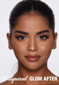 Nahaufnahme eines Gesichts mit glatter, strahlender Haut. Dunkles Haar nach hinten gebunden, definierte Augenbrauen und glänzende nude Lippen vor einem hellen Hintergrund.