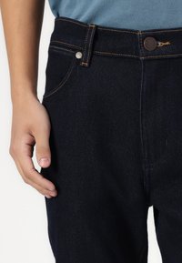 Jeans de denim azul-escuro com corte reto, costura visível, fecho de botão e bolsos laterais. Mão pousada na cintura.