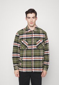 Casaco xadrez verde com riscas brancas, vermelhas e pretas. Apresenta dois bolsos no peito, fecho frontal com zip e gola. Tecido suave e texturizado.