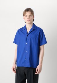 Camisa azul de manga curta em tecido leve, com colarinho pontudo, fecho de botões e um bolso de remendo no lado esquerdo.