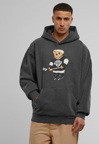Tumši pelēks hoodie, kas izgatavots no mīkstas kokvilnas, ar lāča grafiku sporta ekipējumā, priekšējo kabatu un ribotām aprocēm un apakšmalu.