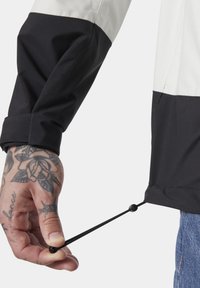 Schwarz-weißer, wasserdichter Jackenärmeln mit elastischem Zugregler. Sichtbare Handgriffe, die ein Tattoo am Handgelenk zur Schau stellen.