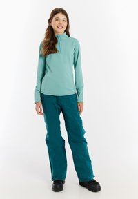 Zipsovaná, dlhá rukávová mikina v mätovej farbe spárovaná s azúrovými snežnými nohavicami a čiernymi čižmami, vyrobená z hladkej, elastickej látky.