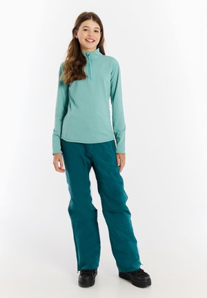 Een geprinte, lange mouw, mintgroene top met rits, gecombineerd met teal sneeuwbroeken en zwarte laarzen, gemaakt van een soepele, rekbare stof.
