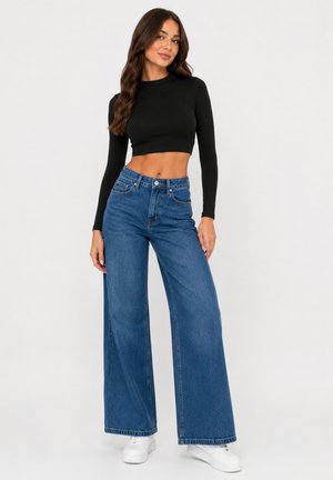 Woman wearing a black long-sleeve crop top, wide-leg blue jeans, and white sneakers, standing against a plain light background.